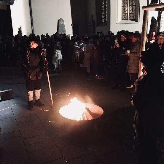 Eine Menschenmenge versammelt sich um ein Lagerfeuer im Freien, mit einer Person daneben, die einen Stock hält. Die Leute sind warm gekleidet und einige haben Mützen. Der Bereich wird vom Feuer beleuchtet.