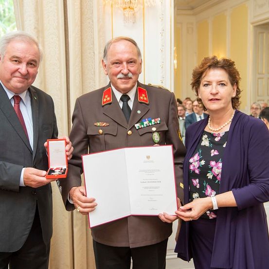 Ein Mann in Uniform, eine Frau in einer Bluse mit Blumenmuster und ein Mann im Anzug präsentieren ein Zertifikat an das Publikum.