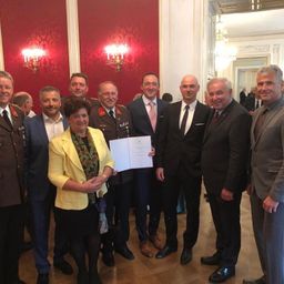 Eine Gruppe formell gekleideter Personen steht in einem Raum und lächelt für ein Foto. Sie halten ein Dokument in der Hand und sind von roten Wänden und Kronleuchtern umgeben.