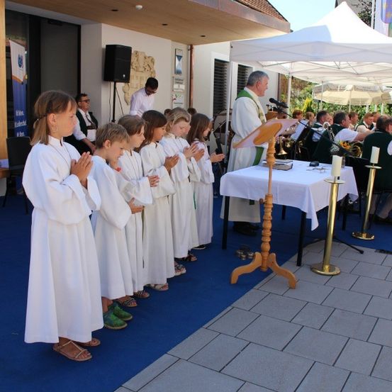 Eine Gruppe von Kindern in weißen Gewändern betet in einer Kirche, während ein Priester an der Kanzel steht. Dahinter spielt eine Band Musikinstrumente.