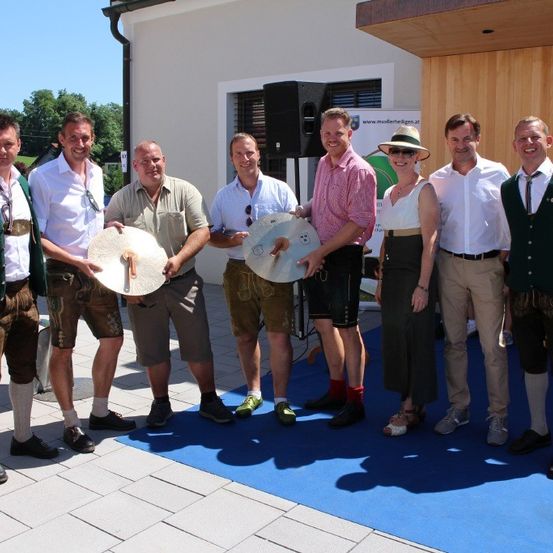 Sieben Männer in traditioneller bayerischer Tracht stehen auf einem blauen Teppich. Zwei halten runde Objekte. Eine Frau in einem Hut steht neben einem Mann in einem rosa Hemd. Ein Lautsprecher und ein Gebäude sind im Hintergrund.