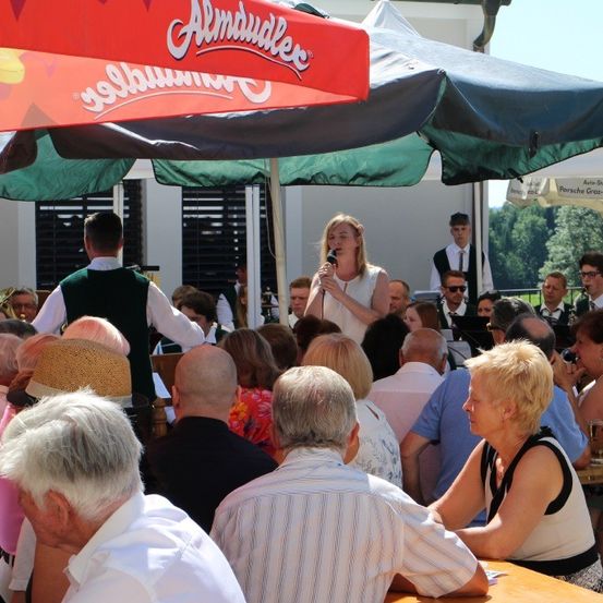 Eine Versammlung im Freien mit mehreren Personen unter Schirmen, eine Frau singt in ein Mikrofon mit einer Band dahinter und ein Schirm mit dem Wort Almdudler.