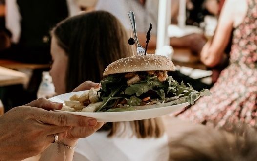 Eine Person hält einen Teller mit einem Burger und Pommes. Eine Gabel ist auf dem Burger. Eine Frau schaut auf den Teller.