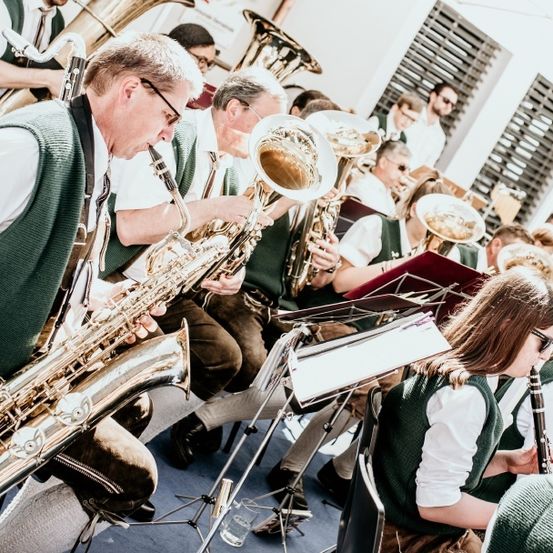 Eine Band spielt auf Blech- und Holzblasinstrumenten; Musiker tragen grüne Westen, und eine Frau spielt Klarinette.