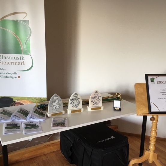 Ein Tisch zeigt Auszeichnungen und Zertifikate auf einer Bühne. Dahinter eine weiße Wand mit einem Banner, das 'Blasmusik Steiermark' und anderen Texten beschriftet ist. Der Raum hat Holzböden.