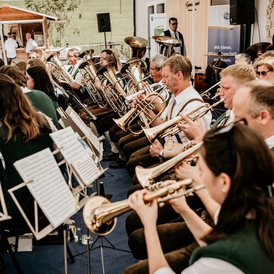 Ein Blechbläserensemble spielt auf einer Bühne mit verschiedenen Blechblasinstrumenten, begleitet von einem Mann in Anzug und Krawatte. Zuhörer sitzen vor ihnen, und ein Banner ist hinter der Band angebracht.