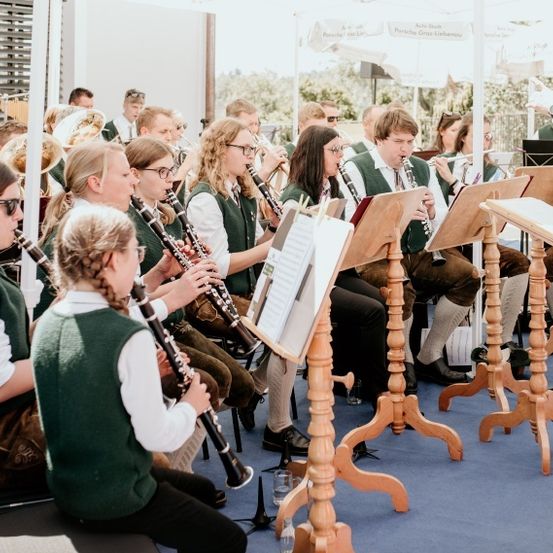 Eine Gruppe junger Musiker in grünen Uniformen und mit Brillen spielt Instrumente bei einem Open-Air-Konzert. Sie sitzen auf Stühlen mit Notenständern. Ein Dirigent leitet die Aufführung.