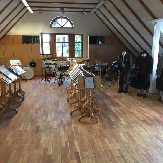 Ein Dachbodenraum mit Holzböden ist mit Musikinstrumenten gefüllt, darunter Trommeln, Becken und Notenständer. Mannequins in formeller Kleidung stehen in der Nähe eines Fensters.