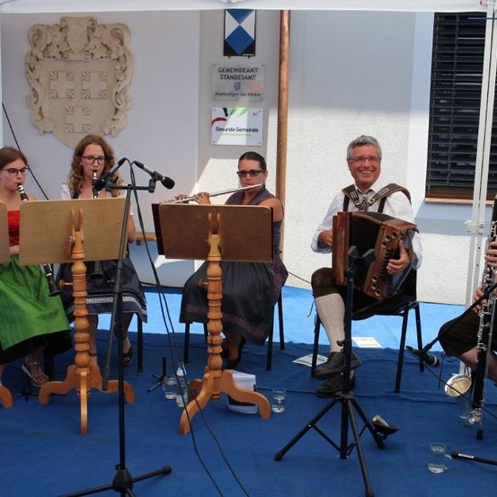 Fünf Musiker spielen auf einer Bühne mit einem blauen Teppich. Zwei Frauen spielen Klarinette, eine spielt Flöte und ein Mann spielt Akkordeon. Eine weitere Person spielt in der rechten Ecke Klarinette. Sie haben Mikrofone auf Ständern vor sich, und es gibt Flaschen und Gläser auf der Bühne.