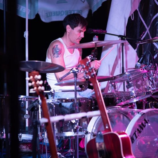 Ein Mann mit einer Tätowierung am Arm spielt die Drums, positioniert unter einem Zelt mit einem Schirm. Ein Mikrofon steht vor ihm, und die Drums sind vor einem weißen Hintergrund aufgebaut.