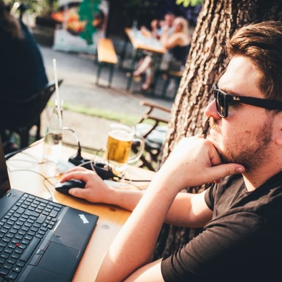 Ein Mann sitzt an einem Tisch in einem Café im Freien und arbeitet an einem Laptop mit einem Bier neben ihm. Er trägt eine Sonnenbrille und hat seine Hand auf dem Kinn, scheinbar in Gedanken versunken.