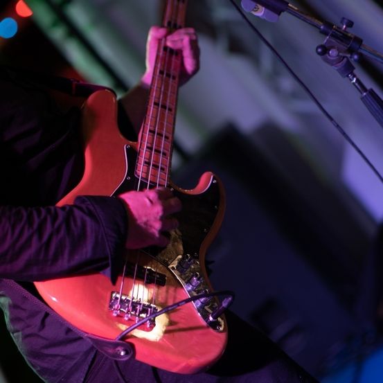 Eine Person spielt eine rote Bassgitarre auf der Bühne unter lila Lichtern. Der Bass hat einen schwarzen Plektrumhalter und weiße Saiten.