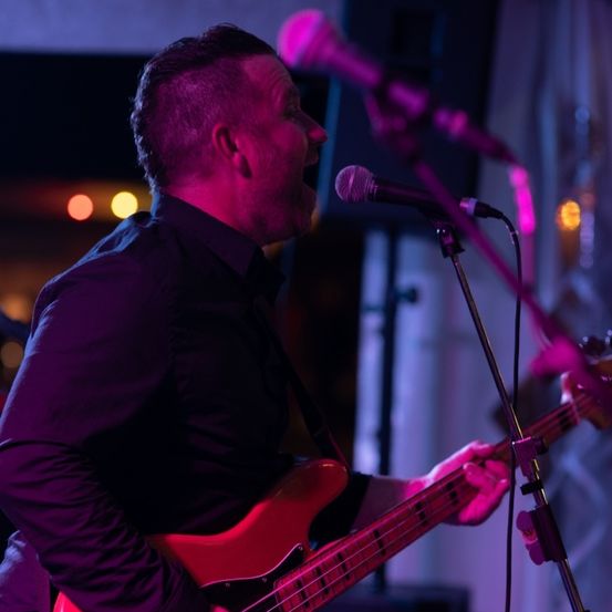 Ein Mann spielt eine rote Bassgitarre auf der Bühne und singt in ein Mikrofon, wobei ein lila Licht die Szene erhellt.