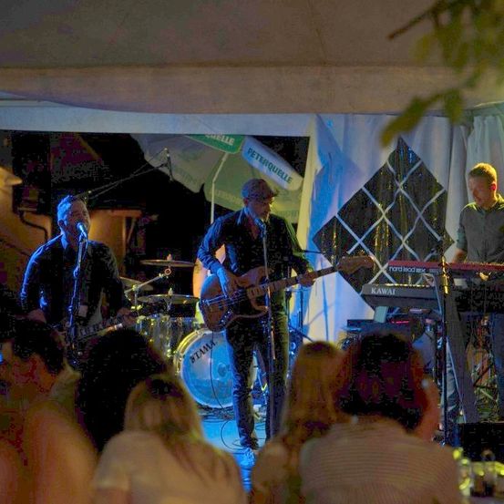 Eine Band spielt bei Nacht auf der Bühne. Ein Mann singt, ein anderer spielt Gitarre und ein anderer spielt Keyboard. Das Publikum beobachtet die Aufführung.