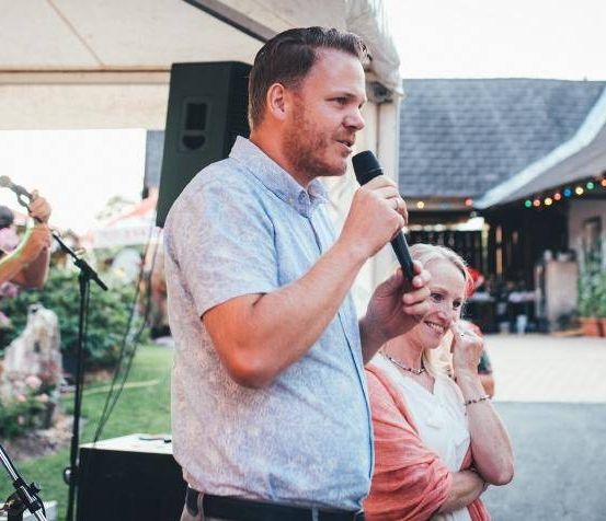 Ein Mann spricht in ein Mikrofon bei einer Veranstaltung im Freien, eine Frau und ein anderer Mann stehen in der Nähe. Ein Haus und ein Garten sind im Hintergrund zu sehen.