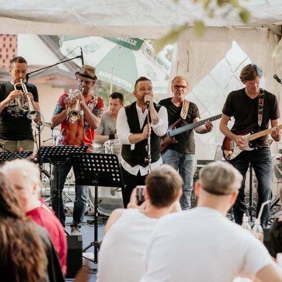 Eine Band tritt auf einer Bühne vor einem großen Publikum auf. Die Band besteht aus einem Trompeter, einem Sänger und Gitarristen. Das Publikum genießt die Aufführung.