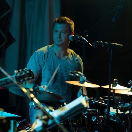 Ein Mann spielt Schlagzeug auf der Bühne bei einem Konzert, mit einem Mikrofon vor ihm und verschiedenen Instrumenten und Ausrüstung um ihn herum.