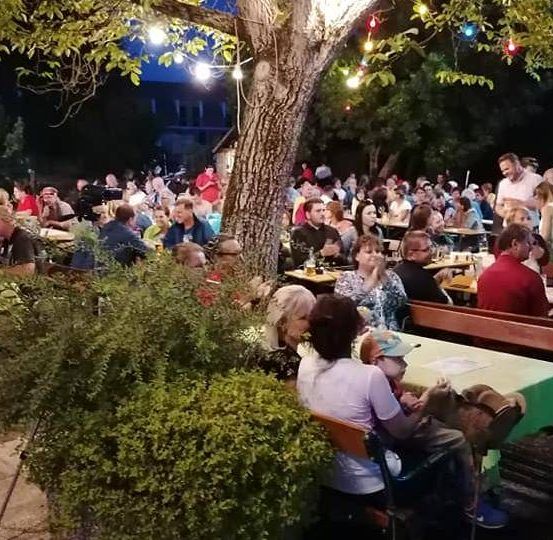 Bei Nacht versammelt sich eine große Menschenmenge in einem Garten im Freien und genießt Getränke und Speisen an Tischen, die mit grünem Tuch bedeckt sind. Der Bereich ist mit bunten Lichtern beleuchtet und mit hängenden Dekorationen geschmückt.