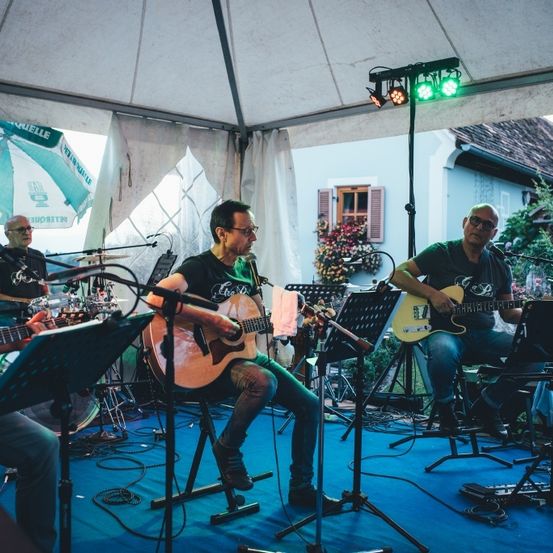 Eine Gruppe Musiker tritt unter einem Zelt mit Mikrofonen und Gitarren auf. Zwei Musiker sitzen auf Hockern, während ein anderer auf einem Hocker steht. Im Hintergrund befinden sich Lichter und ein Schlagzeug.