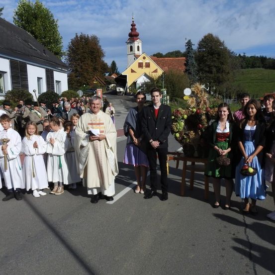 Ein Priester in weißen Gewändern führt eine Gruppe von Kindern in weißen Gewändern an einem sonnigen Tag vor einer Kirche an.