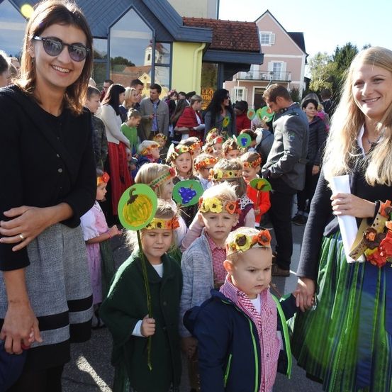 Zwei Frauen stehen mit Kindern in einem festlichen Umzug, tragen Blumenkränze, während Zuschauer und Gebäude im Hintergrund sind.