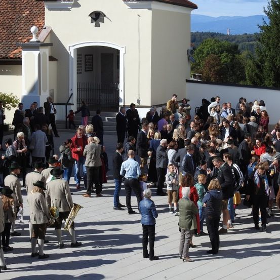 Eine große Menschenmenge versammelt sich vor einer Kirche, Musiker spielen Blechblasinstrumente, einige in traditioneller Kleidung. Das Gebäude hat ein Ziegeldach und eine gerahmte Tür, mit einer Kulisse aus Grün und Bergen.