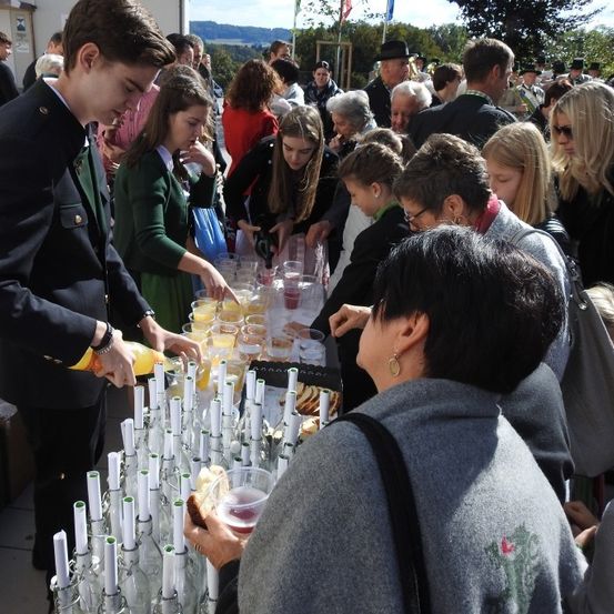 Eine große Gruppe von Menschen, wahrscheinlich bei einer Veranstaltung im Freien, versammelt sich um einen Tisch voller verschiedener Getränke und Speisen. Eine Person in Uniform gießt Getränke in Gläser.