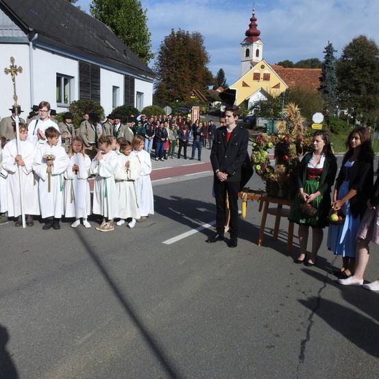 Eine Gruppe von Menschen, einige in weißen Roben, steht auf einer Straße. Ein Mann in einem schwarzen Anzug spricht zu ihnen. In der Nähe steht ein kleiner Tisch mit Früchten, und mehrere Frauen in traditioneller Kleidung stehen dort.