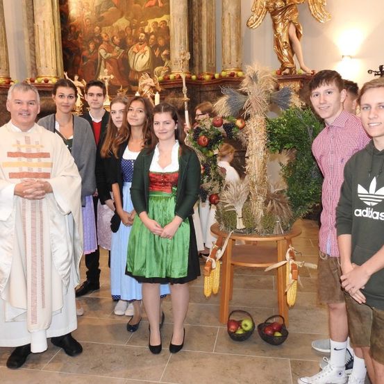Eine Gruppe von Menschen steht lächelnd in einer Kirche. Ein Priester in einer weißen Robe steht neben ihnen. Eine Frau in einer traditionellen bayerischen Tracht steht vor einem dekorativen Tisch mit Äpfeln. Eine goldene Statue steht hinter ihnen.