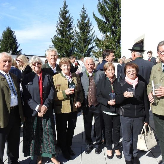 Eine Gruppe von Menschen, sowohl Männer als auch Frauen, die formell gekleidet sind, stehen zusammen vor einem Gebäude mit Tannen im Hintergrund. Sie halten Gläser in ihren Händen.