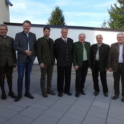 Eine Gruppe von sieben Männern steht draußen in formeller Kleidung. Sie lächeln und scheinen für ein Foto zu posieren. Der Hintergrund zeigt eine weiße Wand mit Bäumen.