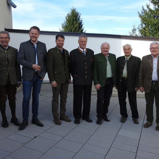 Eine Gruppe von sieben Männern steht draußen in formeller Kleidung. Sie lächeln und scheinen für ein Foto zu posieren. Der Hintergrund zeigt eine weiße Wand mit Bäumen.