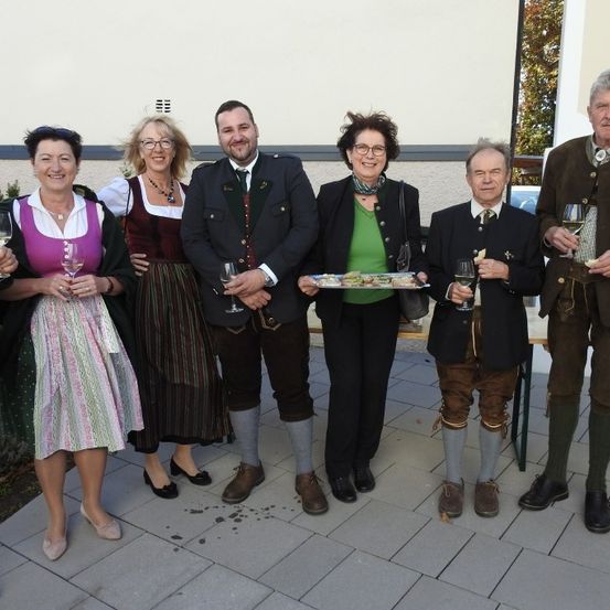 Eine Gruppe von Menschen in traditioneller Kleidung steht zusammen auf einem gefliesten Boden, hält Weingläser in den Händen und lächelt für ein Foto.