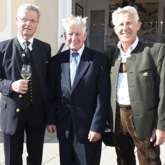 Drei ältere Männer in formeller Kleidung stehen zusammen auf einem Bürgersteig. Einer hält ein Weinglas, und sie alle scheinen für ein Foto zu lächeln. Hinter ihnen steht ein Gebäude mit einem schwarzen Zaun.