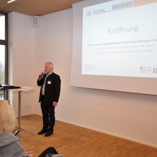 Ein Mann steht vor einer Projektionswand und spricht in ein Mikrofon. Eine Frau sitzt vor ihm. Die Leinwand zeigt eine Folie mit dem Wort 'Eröffnung' in großen Buchstaben.