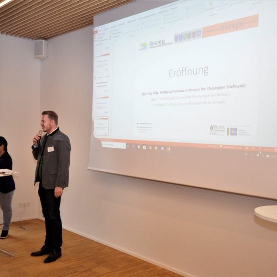 Ein Mann hält einen Vortrag auf einer Konferenz vor einer Projektionsleinwand, die eine Folie mit dem Titel 'Eroffnung' zeigt. Eine andere Person steht in der Nähe und hört zu.