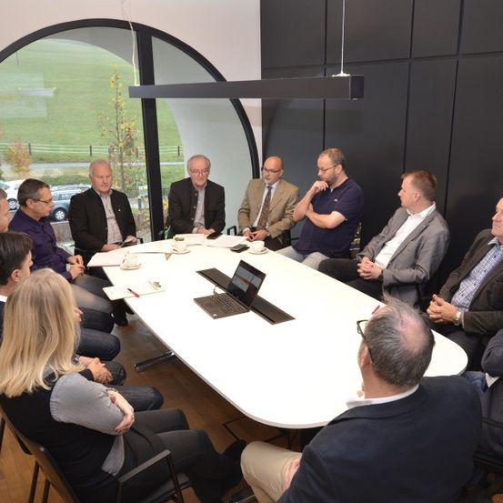 Eine Gruppe von Menschen sitzt in einem modernen Besprechungsraum um einen Tisch herum. Einige führen Gespräche, während andere sich auf den Laptop auf dem Tisch konzentrieren. Der Raum hat große Fenster und Blick auf ein üppiges grünes Feld.