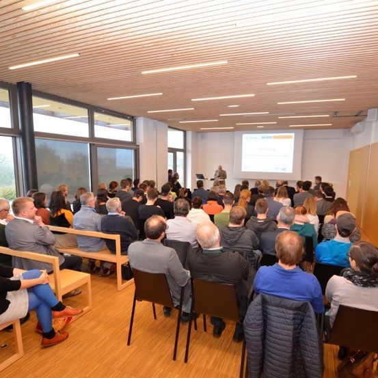 Ein Konferenzraum mit sitzenden Teilnehmern, einer Leinwand an der Wand und einem Mann, der an einem Podium steht.