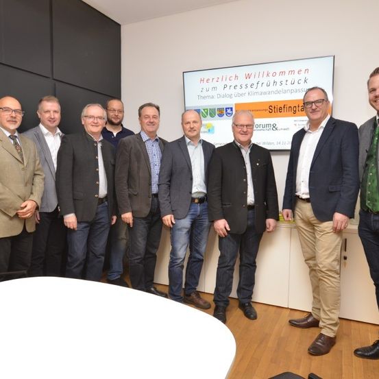 Eine Gruppe von Männern in formeller Kleidung posiert für ein Foto vor einem großen Schild, das 'Herzlich Willkommen zum Pressefruhstuck. Thema: Dialog uber Klimawandelanpassung. Stiftung.' steht.