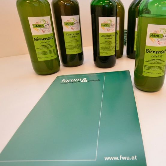 Mehrere grüne Flaschen mit Etiketten und eine grüne Mappe mit dem Text 'www.fwu.at' sind auf einem weißen Tisch ausgestellt.