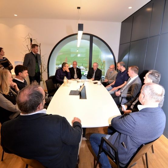 Eine Gruppe von Fachleuten sitzt um einen langen Tisch in einem modernen Besprechungsraum. Ein Laptop und einige Papiere liegen auf dem Tisch. Der Raum hat große Fenster und eine geschwungene Wand.