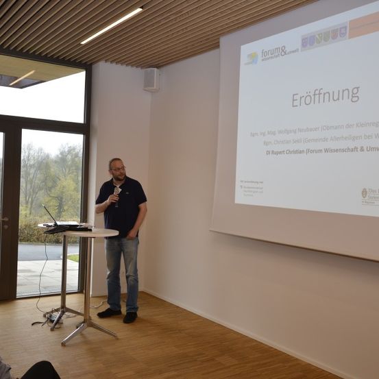 Ein Mann gibt einen Vortrag in einem Konferenzraum. Die Beamerfolie hinter ihm zeigt eine Folie mit der Überschrift 'Eröffnung'. Der Raum hat Glastüren und Fenster, einen Holzboden und eine Glastür, die nach außen führt.