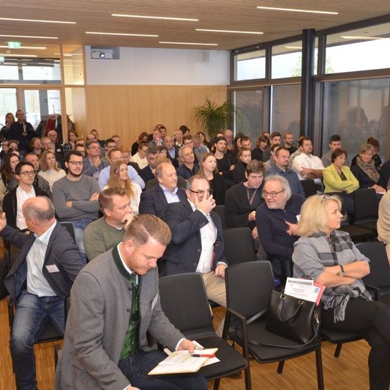 Eine große Gruppe von Menschen sitzt in einem Konferenzsaal und trägt formelle Kleidung. Einige unterhalten sich, während andere aufmerksam zuhören. Eine Frau hält ein Stück Papier in der Hand und scheint Notizen zu machen.