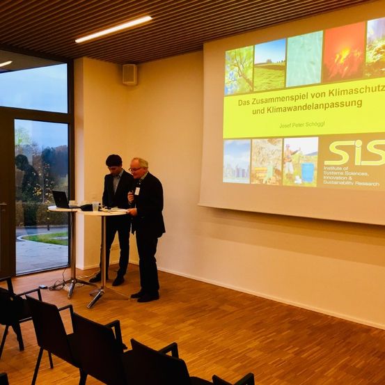 Zwei Männer stehen in einem Raum neben einem Tisch mit einem Laptop und blicken auf eine Projektionsleinwand mit einer Präsentation mit dem Titel 'Das Zusammenspiel von Klimaschutz und Klimawandelanpassung.' Stühle sind davor aufgestellt.