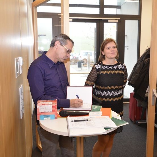 Ein Mann mit Brille unterschreibt ein Dokument, während eine Frau neben ihm steht und lächelt. Sie befinden sich in einem Raum mit einem Tisch, auf dem Bücher, ein Notizbuch, ein Mikrofon und eine Handtasche stehen. Hinter ihnen ist eine Glastür mit einem Baum im Freien sichtbar.