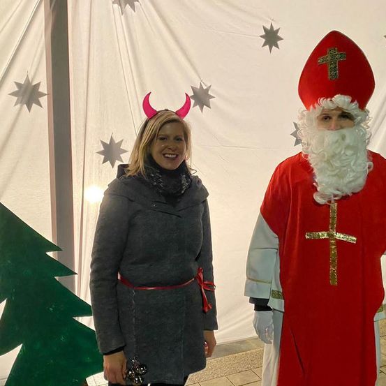 Eine lächelnde Frau in einem grauen Mantel und einer roten Teufelshorn-Kopfbedeckung posiert neben einem Mann, der als Weihnachtsmann verkleidet ist, vor einem Weihnachtsbaum.