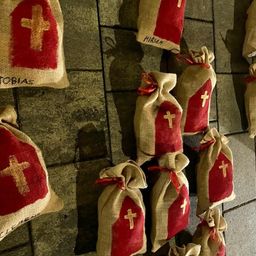 Eine Reihe von Jutebeuteln mit roten Kreuzen und goldenen Kreuzdesigns ist an einer Wand befestigt. Einige Beutel haben Namen wie Tobias und Mirah.