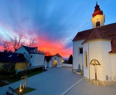 Ein Sonnenuntergang über einer Kirche mit einem roten Dach, einem Kreuz an der Wand und einem Weg, der zum Eingang führt. Bäume und Gebäude im Hintergrund.