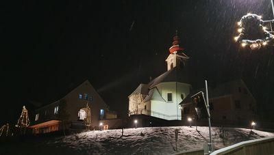 Eine Kirche mit einem Kirchturm steht beleuchtet bei Nacht auf einem verschneiten Hügel, umgeben von Weihnachtslichtern und Dekorationen.