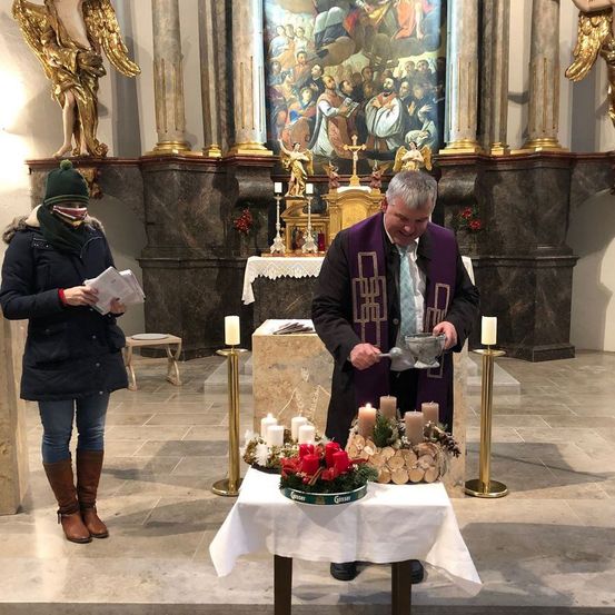 Ein Priester gießt Wasser aus einem Kelch in ein Becken auf einem Tisch mit brennenden Kerzen und Blumen. Eine Frau steht in der Nähe.
